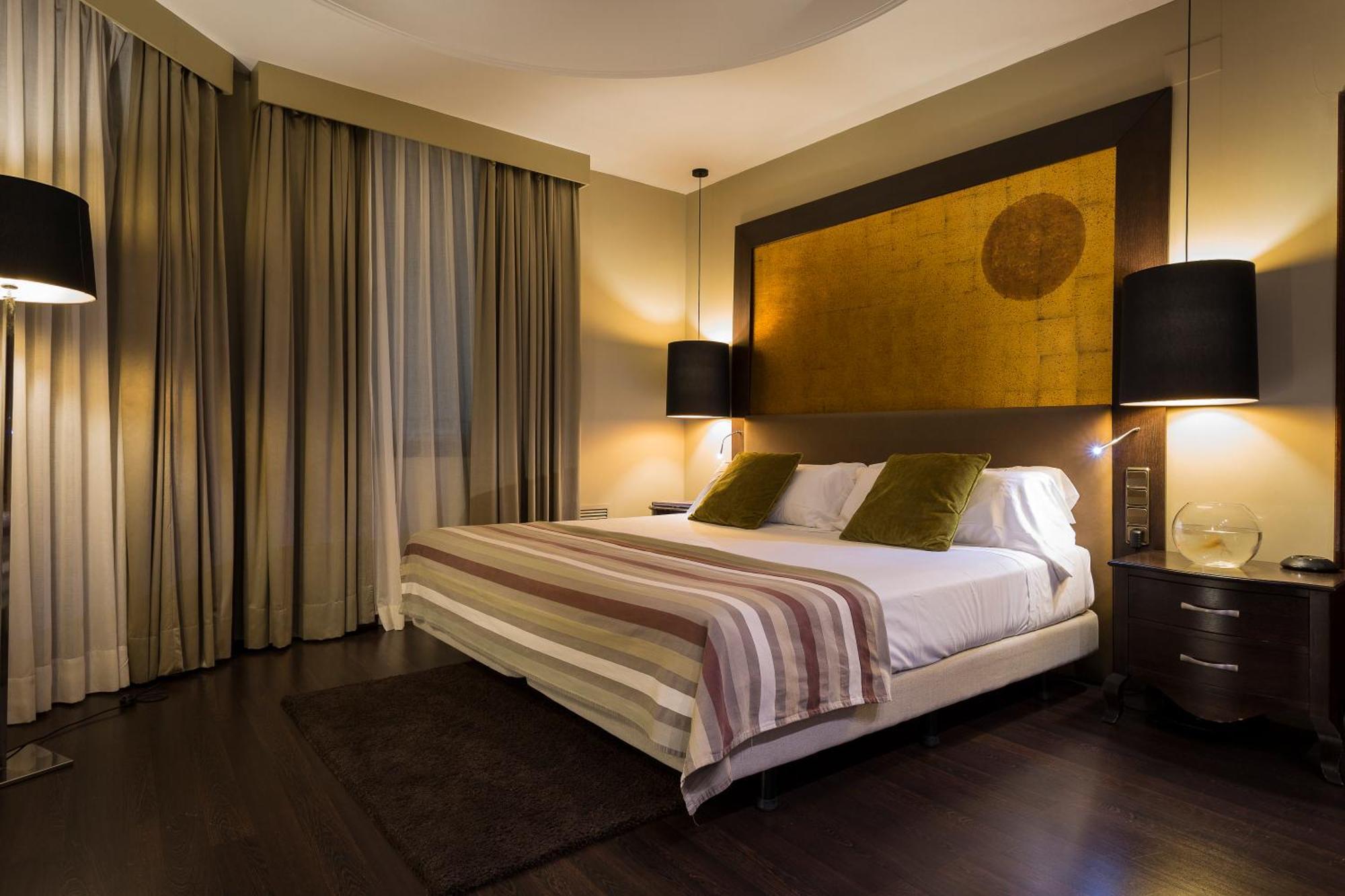Only You Hotel Valencia Ngoại thất bức ảnh The photo shows a modern bedroom that features a large bed with two green accent pillows and a striped bedspread. The room has a warm, neutral color scheme, highlighted by soft lighting from two bedside lamps. The wall behind the bed has an artistic 